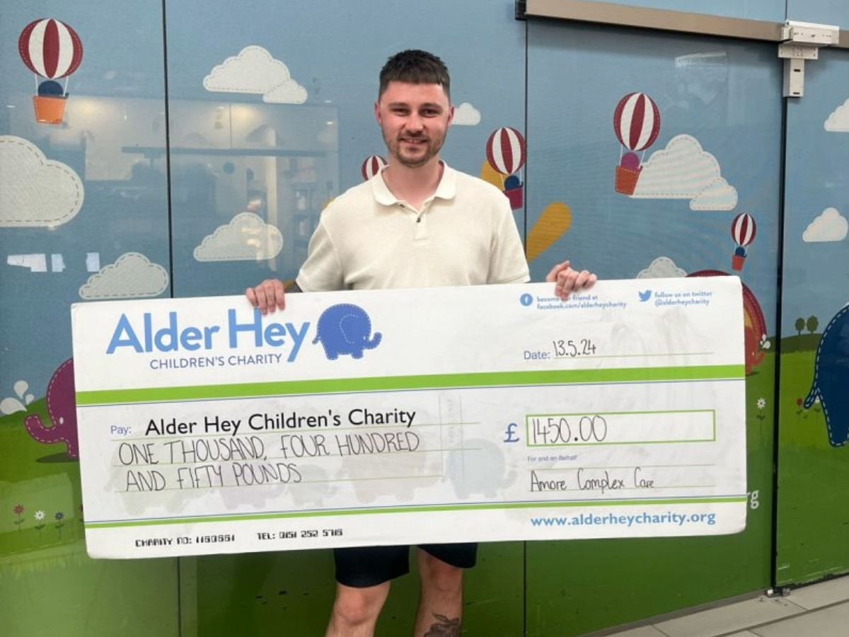 Louis Benfield standing in Alder Hey Children's Charity's offices, holding a check from Amore Complex Care.